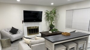 A modern living room with a fireplace, a flat-screen TV mounted above it, a grey sofa with cushions, a small wooden table, and a potted plant. - PickleballShopsFoodies 5Bedrms 2Washers 3Dryers (Montclair)