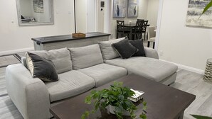A modern living room with a grey sectional sofa, a dark wooden coffee table, and a potted plant. There is a dining area in the background with a white table and chairs. - PickleballShopsFoodies 5Bedrms 2Washers 3Dryers (Montclair)