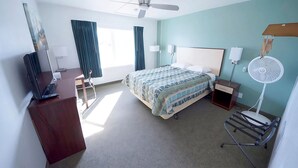 A hotel room with a bed, a desk with a laptop, a standing fan, and a window with curtains. - Rewak Studios (Fairbanks)