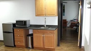 A compact kitchen with wooden cabinets, a microwave, a refrigerator, and a trash bin. - Rewak Studios (Fairbanks)