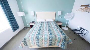 A hotel room with a bed, bedside lamps, a standing fan, a chair, and a window with curtains.