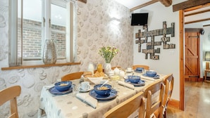 A dining area with a long table set for four, wooden chairs, a brick wall, and a window with blinds. - Nobles Cottage - UK43351 (Whitby)