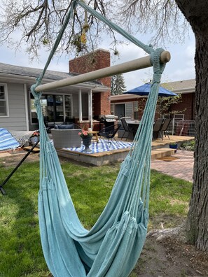 Una hamaca colgada entre dos árboles en un patio trasero con terraza, muebles de exterior y una parrilla.