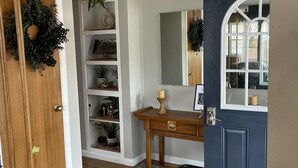 Un pasillo con una puerta de madera, una puerta azul, una pared blanca, un estante con objetos decorativos, una mesita con una vela y una lámpara en el techo.