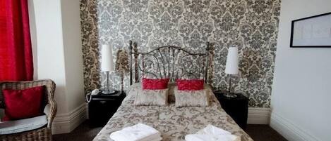 Un dormitorio ordenado con una cama grande, dos mesitas de noche, una silla y papel tapiz estampado.