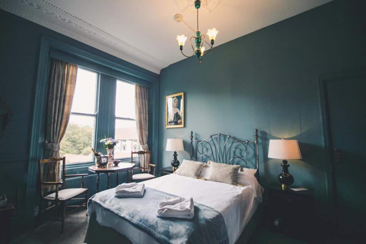 A bedroom with a large bed, two bedside tables with lamps, a chair, a window with curtains, and a chandelier.