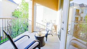 A balcony with wicker chairs, a glass table, and a view of a garden and neighboring buildings. - Luxury condo near Irvine Business & UCI Hospital (Irvine)