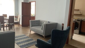 A living room with a grey sofa, a blue armchair, and a dining area with chairs and a table. - Serenity Inn (Quito)