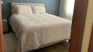 A bedroom with a bed, a nightstand, a wooden shelf, and a window with curtains. - Serenity Inn (Quito)