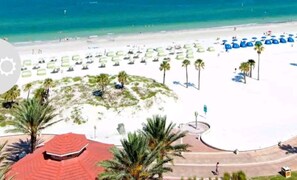 Ein Strand mit Reihen von Sonnenschirmen und Liegestühlen, Palmen und einem Gebäude mit rotem Dach.