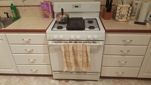 A kitchen with a white stove, oven, and a kettle on the countertop. There are cabinets with gold handles and a roll of paper towels.
