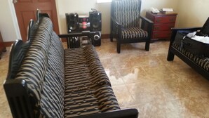 A living room with a patterned sofa, a chair, a small table with speakers, and a piano.