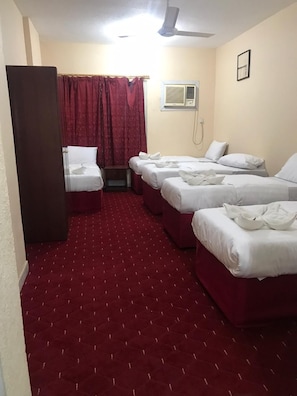 Habitación de hotel con tres camas, ventilador de techo, unidad de aire acondicionado y piso cubierto de alfombra roja.