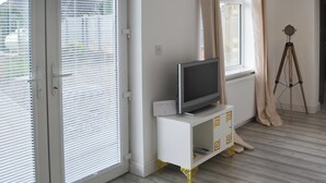 A living room with a TV on a white cabinet, a tripod, and a large window with blinds. - Old Colliery Cottage (Shilbottle, near Alnwick)
