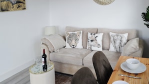 A living room with a beige sofa, three animal-themed cushions, a round mirror, a glass table with a wine bottle, and a side table with a lamp. - Old Colliery Cottage (Shilbottle, near Alnwick)