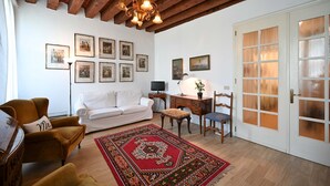 Un salon avec un canapé blanc, un fauteuil moutarde, un bureau en bois et un tapis rouge à motifs.