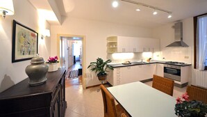 Une cuisine moderne avec des meubles blancs, un coin repas avec une table blanche et des chaises en osier, et un couloir menant à une autre pièce.