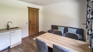 A compact kitchen with a sink and cabinets, a wooden door, a dining table with chairs, and a sofa with cushions.