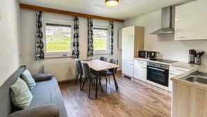 A compact kitchen with a dining table and chairs, a sofa, and a view of a green landscape through the windows.