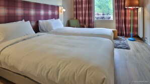 Habitación de hotel con dos camas, una silla, una lámpara y vista a zonas verdes por la ventana.