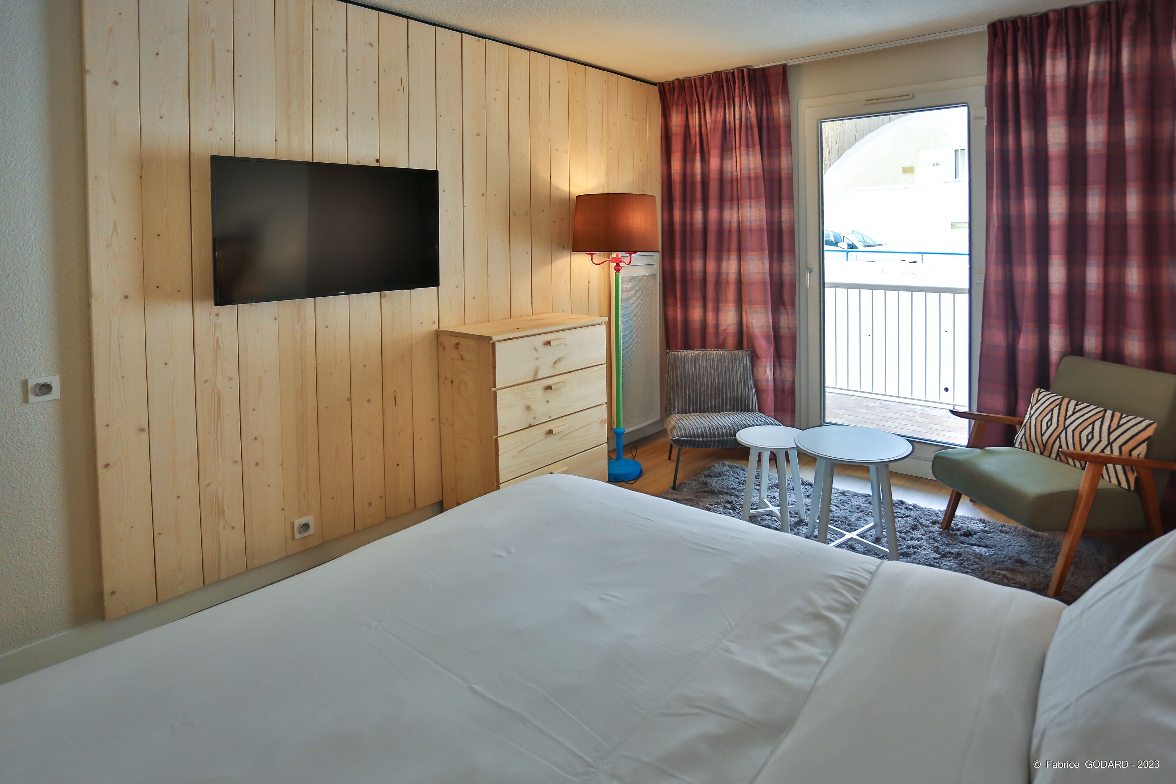 A hotel room with a large bed, a flat-screen TV mounted on a wooden wall, a small table with chairs, and a balcony with curtains.