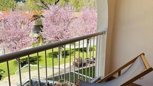 Un balcón con una silla acolchada, una mesita con un libro y una taza, y vistas a flores de cerezo y un edificio al fondo.