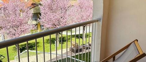 A balcony with a cushioned chair, a small table with a book and a cup, and a view of cherry blossoms and a building in the background.