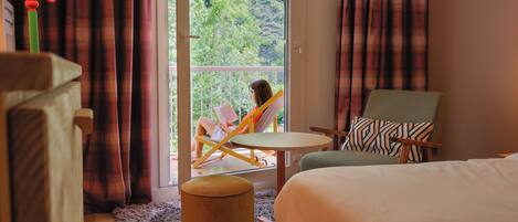 A hotel room with a bed, a chair, a small table, and a view of greenery outside.