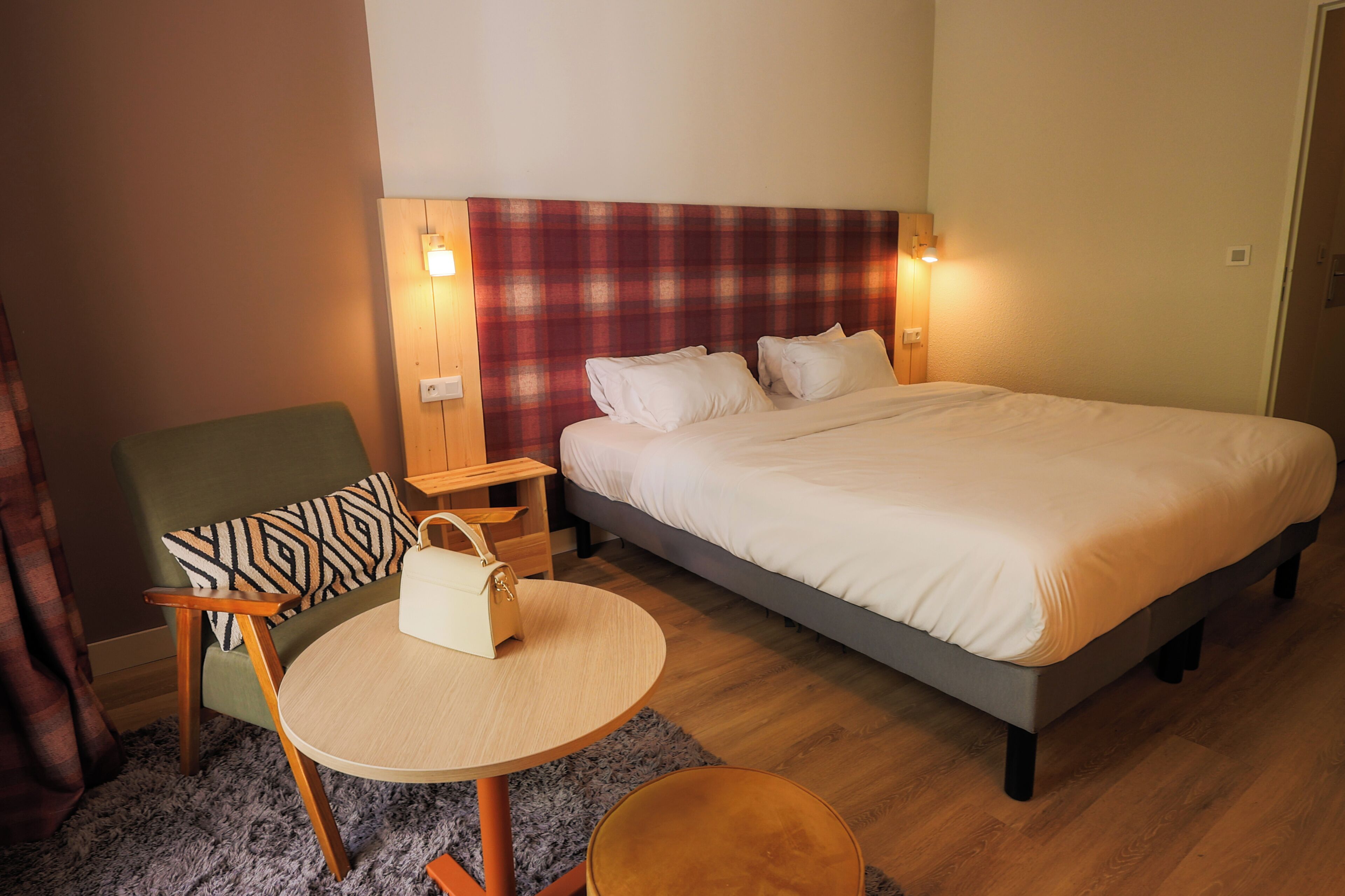 A hotel room with a bed, a chair, a small table, and a nightstand with a lamp.