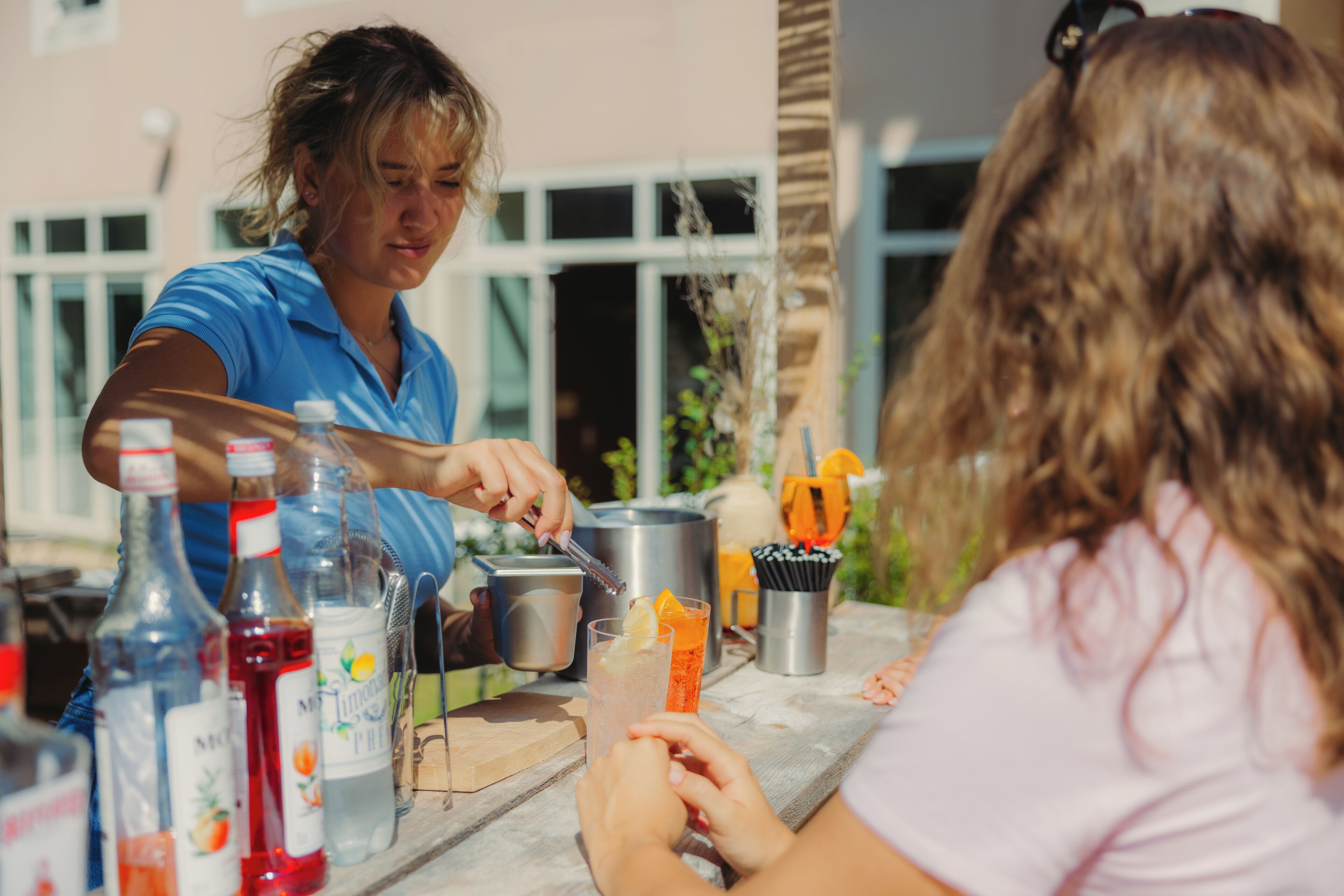 Une personne prépare une boisson à l’extérieur, sur une table, en utilisant différentes bouteilles et un récipient en métal.