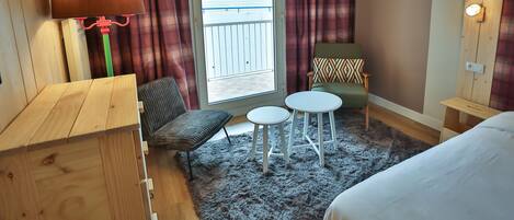 A hotel room with a wooden desk, a green chair, a small round table, a bed, a grey rug, and a window with curtains.