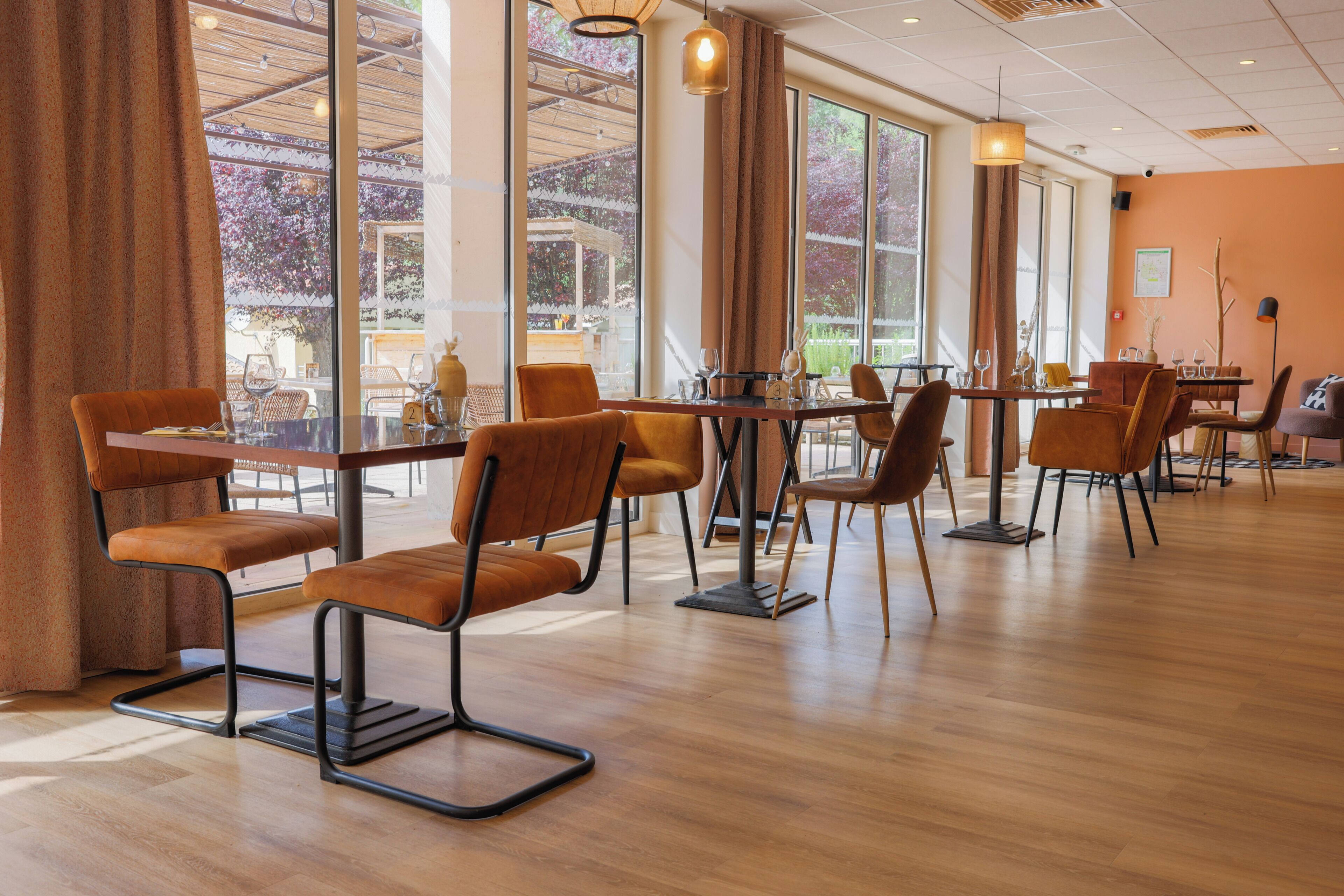Un restaurante con mesas y sillas de madera, ventanas amplias y vistas a árboles.