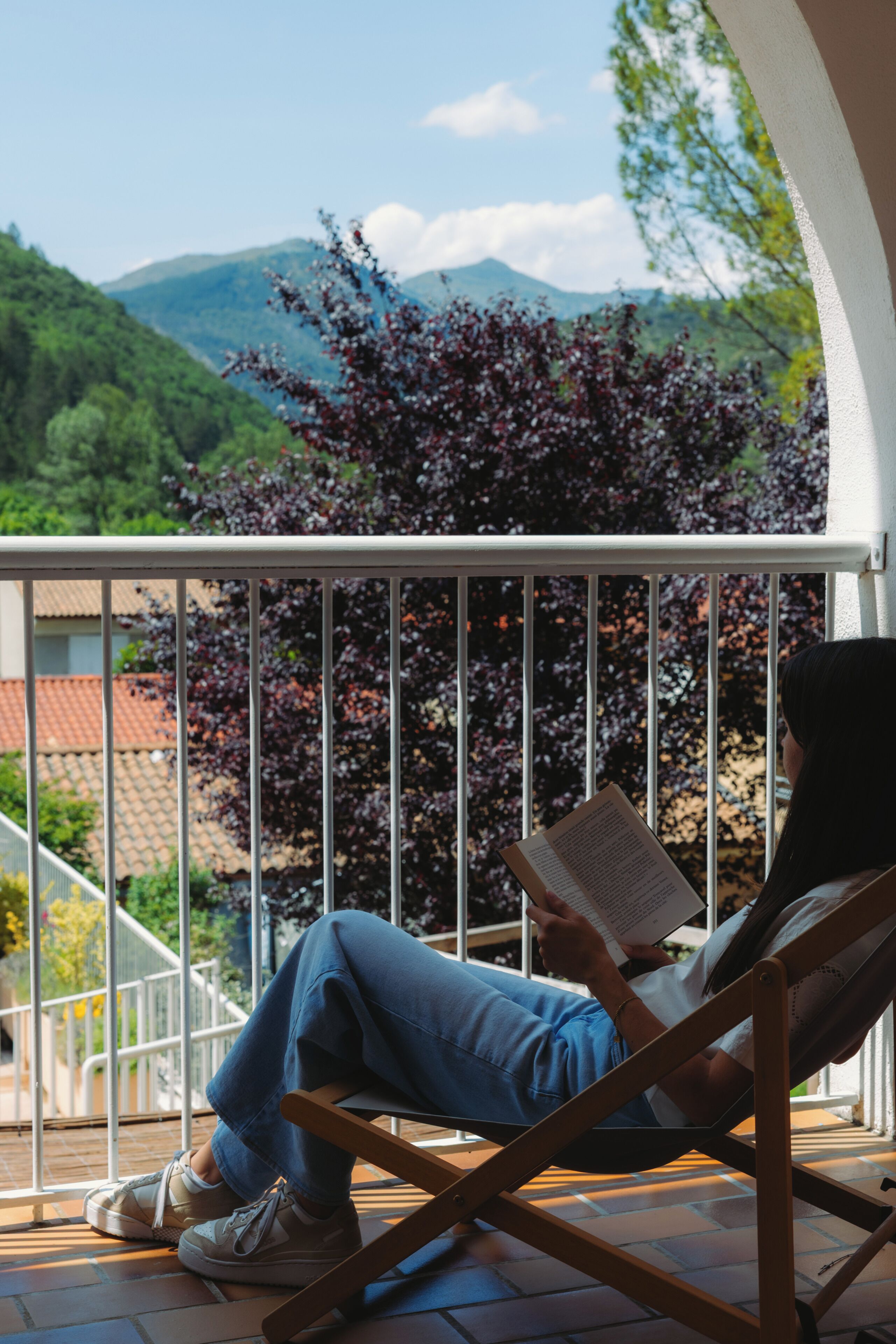 A person reading a book on a balcony with a view of mountains and trees.
