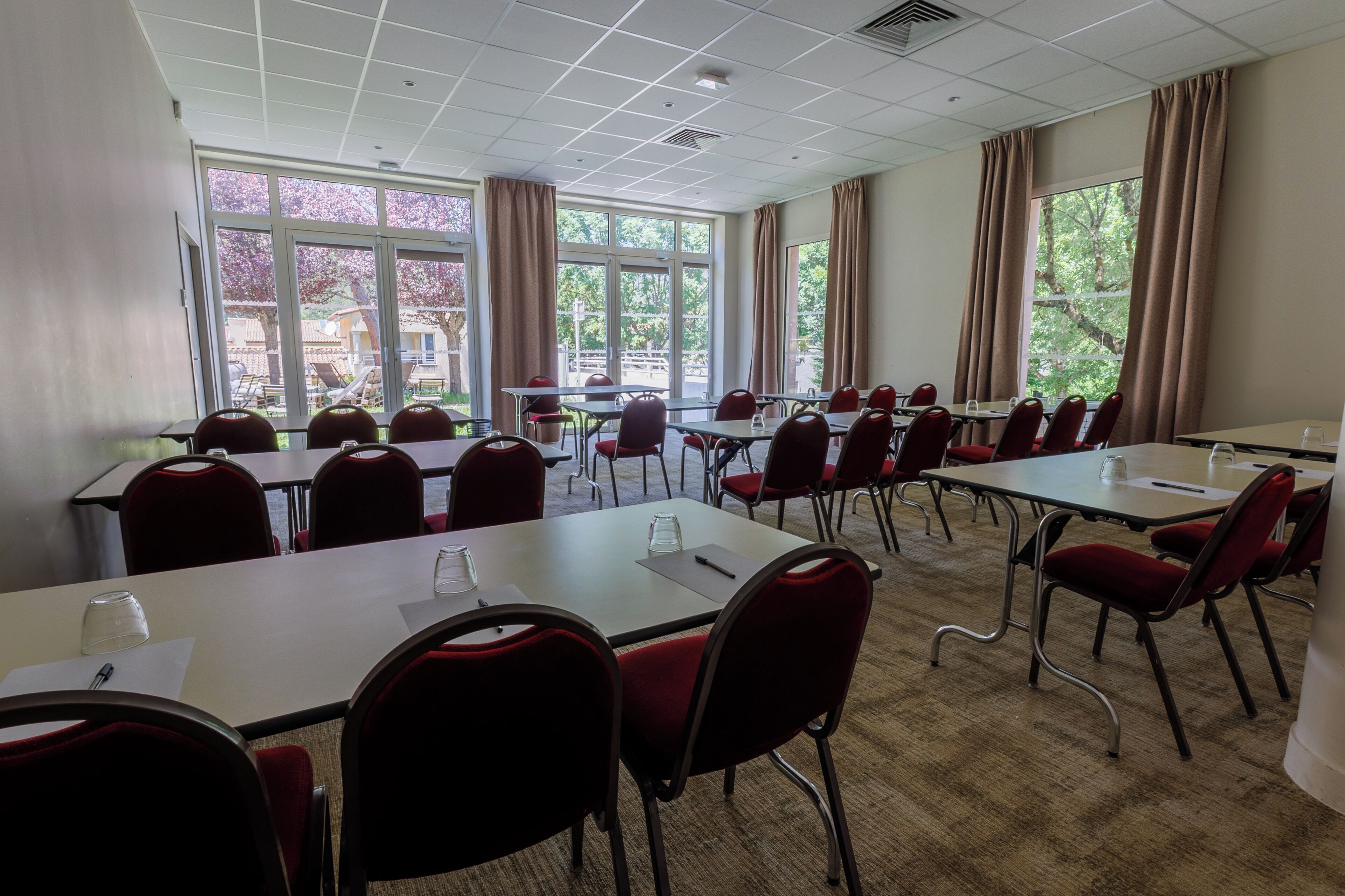 A conference room with multiple tables and chairs, large windows with curtains, and a carpeted floor.
