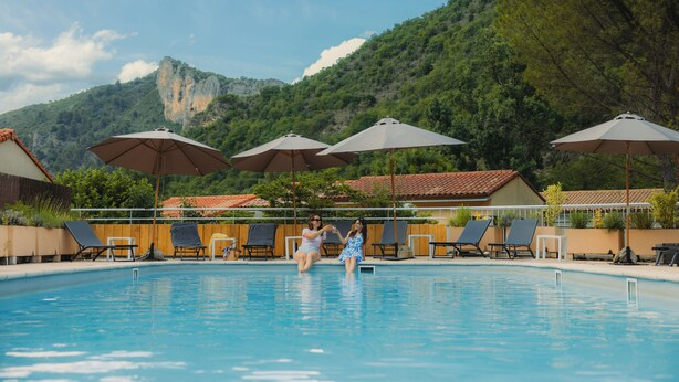 Una piscina con sillas de descanso y sombrillas, rodeada por una valla de madera y una montaña al fondo.