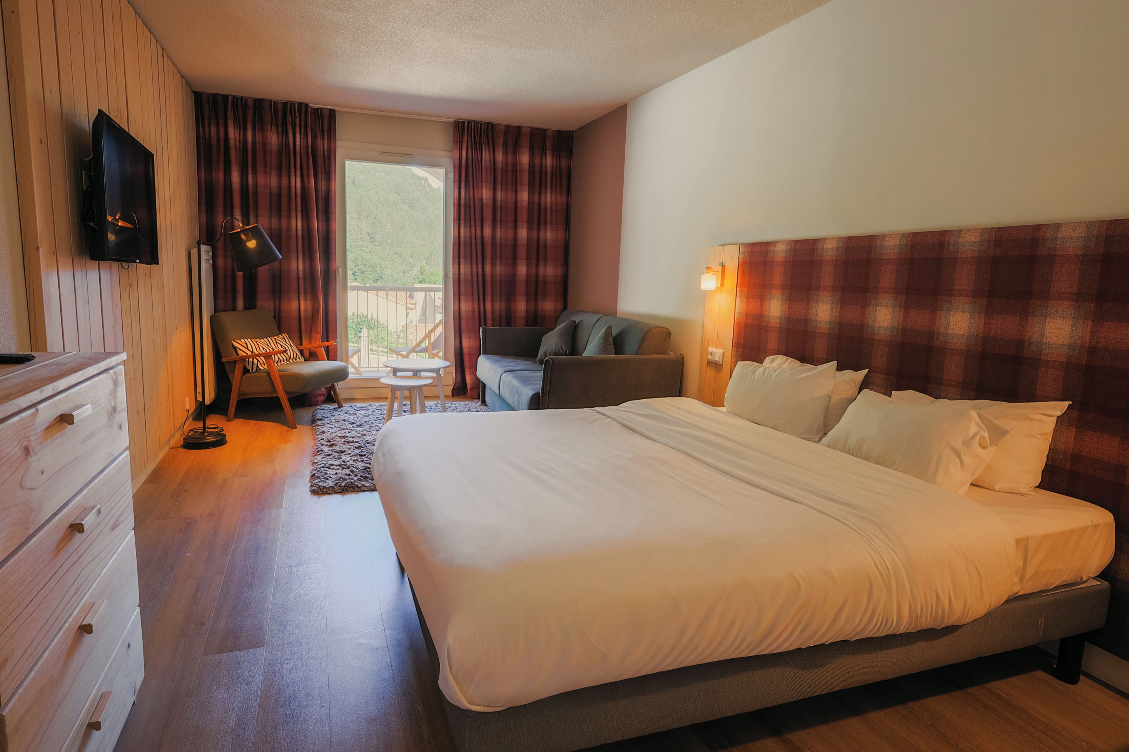 A hotel room with a large bed, a desk, a chair, a TV, and a balcony with curtains.