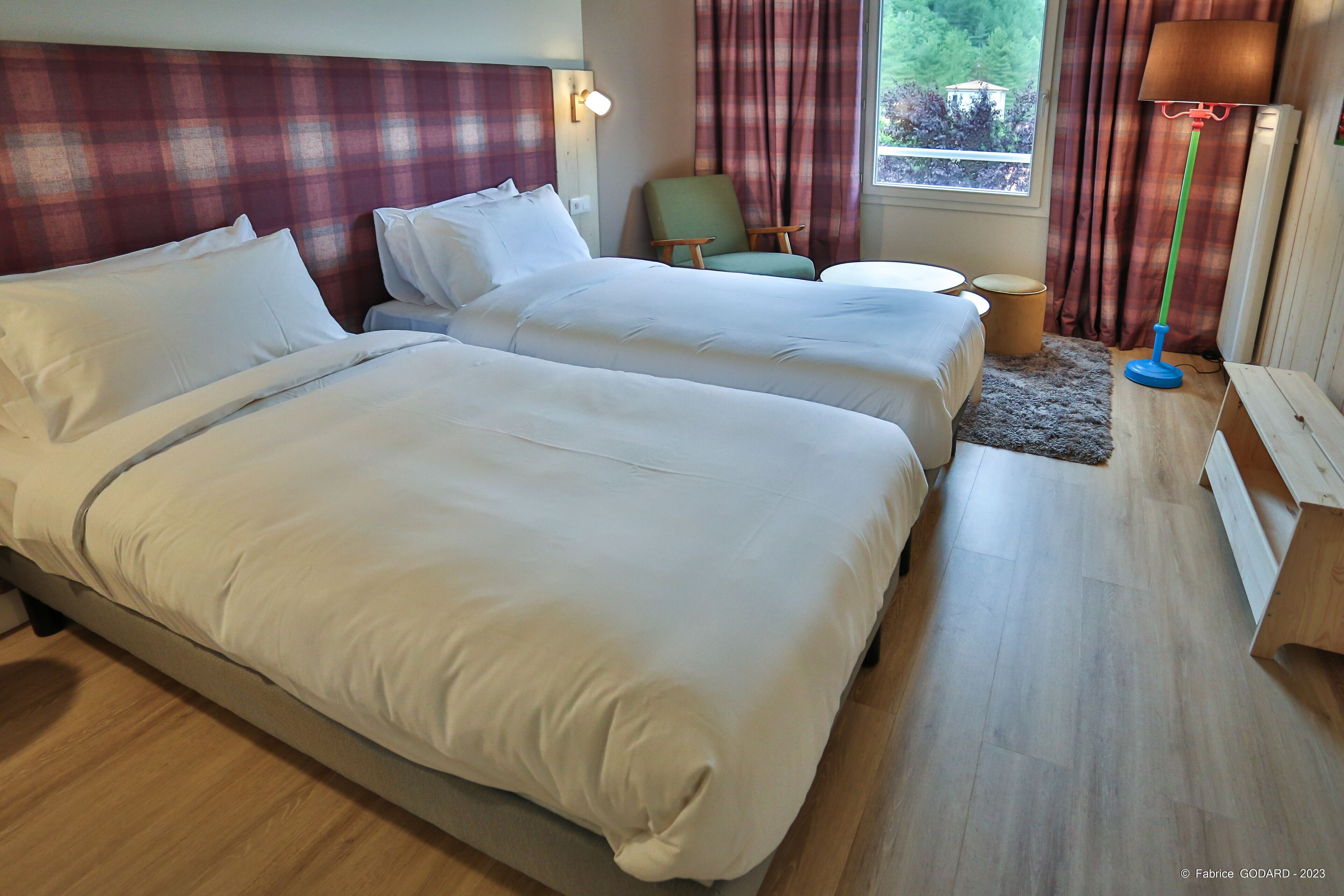 Habitación de hotel con dos camas, una mesita, una silla, una lámpara y vista a zonas verdes.