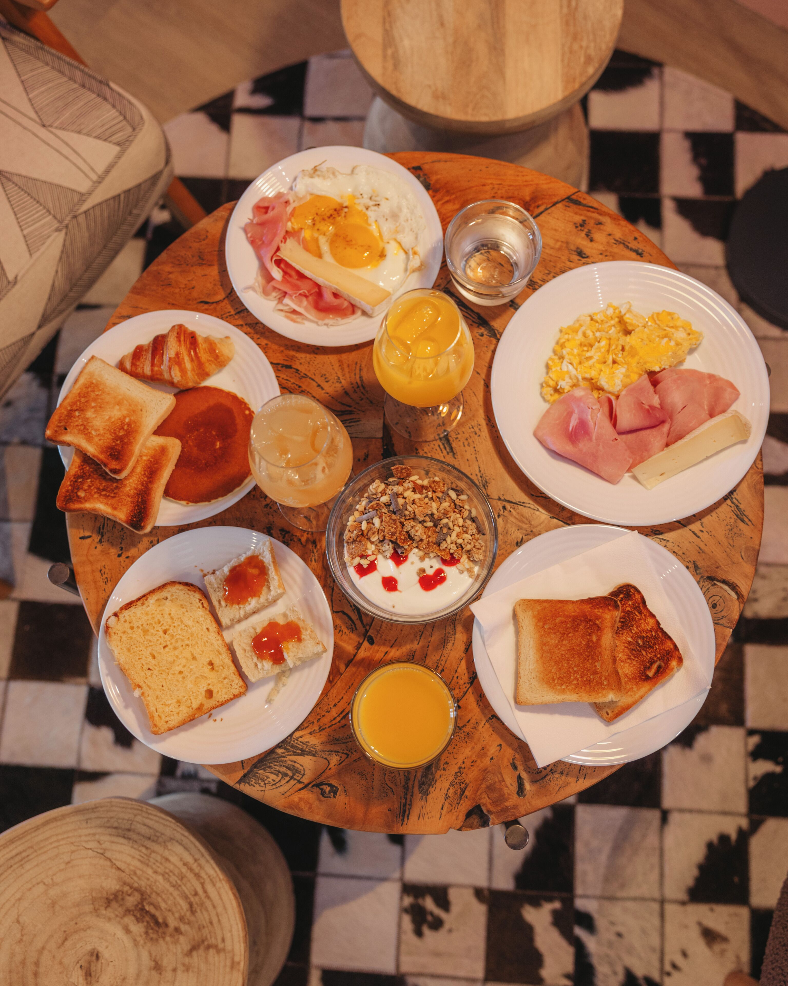 Una mesa de madera redonda con diversos alimentos para el desayuno, como tostadas, huevos, jamón y un vaso de jugo de naranja.