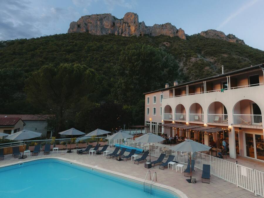 Un área de piscina con sillones reclinables y sombrillas, un edificio con balcones y una montaña al fondo.