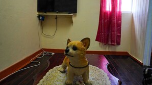 Une petite figurine de chien est posée sur un paillasson orné d’une inscription « BIENVENUE », un téléviseur est fixé au mur et un rideau rouge est partiellement visible.