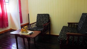 Un salon avec un plancher en bois, une chaise en bois avec un coussin à motifs noir et blanc, un canapé en bois avec un coussin similaire, et une petite table sur laquelle se trouvent une télécommande et un jouet représentant un chien.