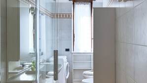 Un baño con cabina de ducha de vidrio, lavadora, inodoro y ventana con cortina.