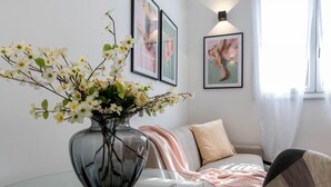 Una sala de estar moderna con una mesa de centro de vidrio, un jarrón con flores blancas, un sofá con una manta rosa y obras de arte enmarcadas en la pared.