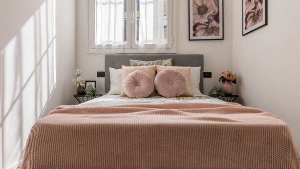 Un dormitorio con cama, dos mesitas de noche, una ventana con cortinas y un cuadro con motivos florales en la pared.