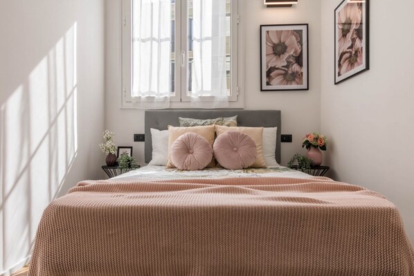 Un dormitorio con cama, dos mesitas de noche, una ventana con cortinas y un cuadro con motivos florales en la pared.