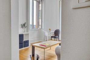 A room with a wooden floor, a desk with a chair, a small table with a magazine, and a window with blinds.