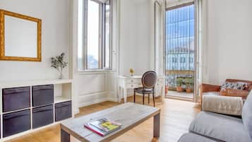 Een moderne woonkamer met een houten salontafel, een bank, een bureau en een stoel. Grote ramen bieden uitzicht op de stad.