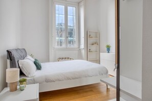 A bedroom with a bed, bedside table, lamp, potted plants, and a window.