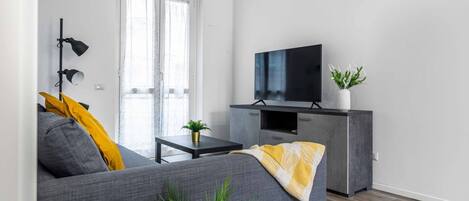 A modern living room with a grey sofa, a flat-screen TV on a cabinet, a small table with a plant, and a window with curtains.
