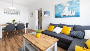 Un salon moderne avec une table basse en bois, un canapé gris agrémenté de coussins jaunes, un coin repas avec une table blanche et des chaises grises, et un tableau représentant des voiliers accroché au mur.